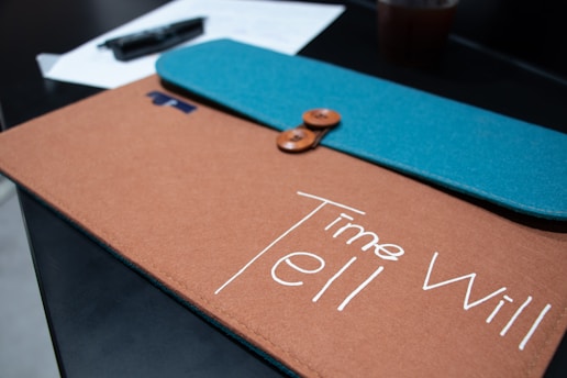 A close-up shot of a brown and teal folder with a button closure, placed on a dark surface. The folder has the phrase 'Time Will Tell' written on it in white letters. In the background, there are a couple of papers and a plastic cup, slightly out of focus.