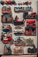 The image features a collection of colorful travel magnets displayed on a vertical surface. These magnets depict various global cities and landmarks such as Frankfurt, Münster, Venice, Barcelona, Berlin, and Cologne. Some are in the shape of famous buildings or monuments, while others display the city's name prominently. The magnets are closely packed, showcasing a variety of designs and bright colors.