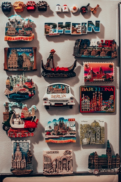 The image features a collection of colorful travel magnets displayed on a vertical surface. These magnets depict various global cities and landmarks such as Frankfurt, Münster, Venice, Barcelona, Berlin, and Cologne. Some are in the shape of famous buildings or monuments, while others display the city's name prominently. The magnets are closely packed, showcasing a variety of designs and bright colors.