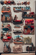 The image features a collection of colorful travel magnets displayed on a vertical surface. These magnets depict various global cities and landmarks such as Frankfurt, M&uuml;nster, Venice, Barcelona, Berlin, and Cologne. Some are in the shape of famous buildings or monuments, while others display the city's name prominently. The magnets are closely packed, showcasing a variety of designs and bright colors.