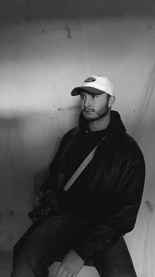 A man is sitting against a plain backdrop, wearing a white baseball cap and a dark jacket. He holds a camera while gazing off to the side, creating a contemplative pose. The image has a vintage, monochrome aesthetic.