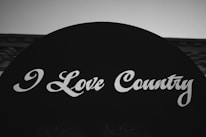 A black and white image featuring a bold sign with the words 'I Love Country' written in a stylized white script font. The background is minimal, focusing attention on the text.