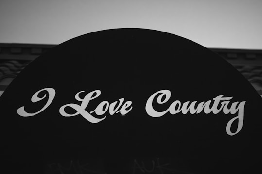 A black and white image featuring a bold sign with the words 'I Love Country' written in a stylized white script font. The background is minimal, focusing attention on the text.