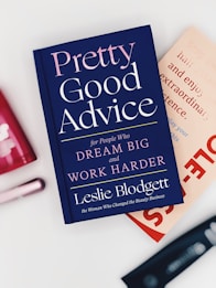 A hardcover book titled 'Pretty Good Advice' is lying on top of another book. The book cover is dark blue with pink and white text, featuring motivational phrases. Around the books are blurred objects, possibly cosmetics or personal care items, placed on a white surface.