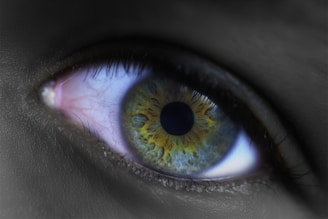 A close-up of a human eye capturing intricate details of the iris. The iris has a vibrant combination of green and yellow hues with a dark pupil in the center. The sclera and surrounding eyelashes are visible, and the image is set in a high contrast with the rest of the skin area appearing in grayscale.