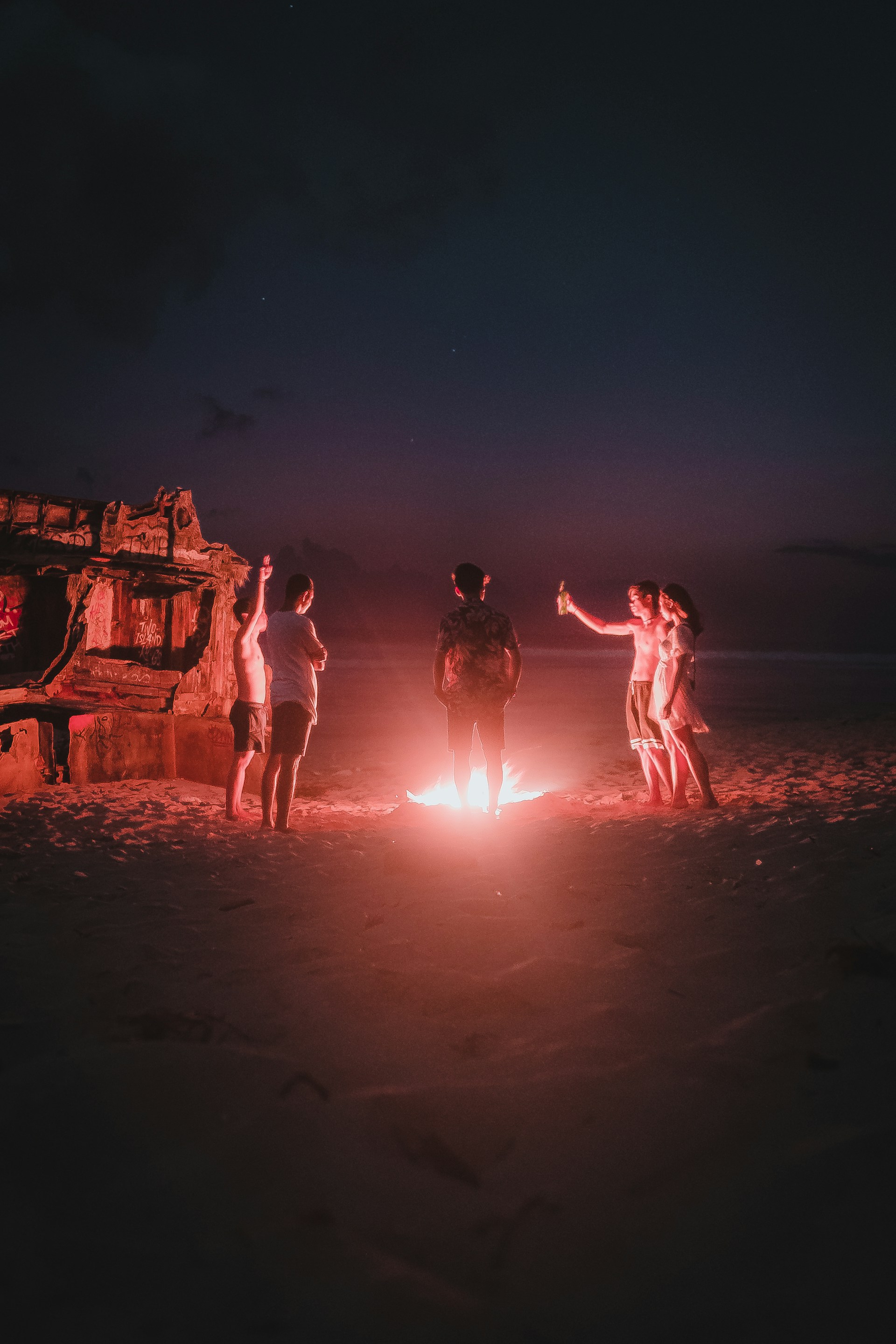 A joyful group of travelers dancing around a bonfire on a beach at dusk, glowing with warm burnt orange light.