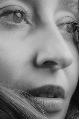 A close-up black and white photograph of a person's face, focusing on the eye, nose, and mouth. The image captures fine details such as eyelashes, skin texture, and strands of hair.