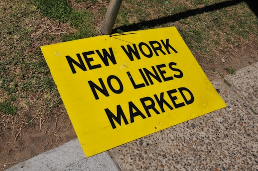 A bright yellow warning sign is placed on a sidewalk and grassy area, displaying the message 'NEW WORK NO LINES MARKED' in bold black letters. The sign leans against a pole, suggesting an ongoing construction or roadwork activity nearby.