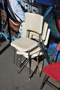 Four stacked vintage chairs with yellowish upholstery and metal legs are placed on an urban street against a colorful wall mural. The mural features ocean elements and bold black lettering. A red metal chair is visible next to the stacked chairs.