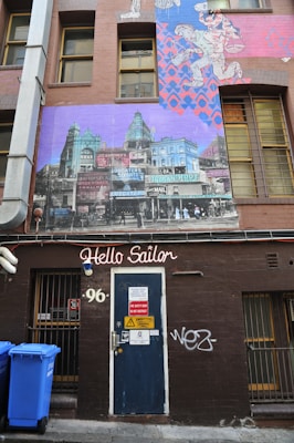 A brown building facade features a large mural with historical architecture in vibrant colors. Above a dark blue door labeled 'Hello Sailor' with assorted warning signs, there are blue and pink street art designs. Two blue trash bins are positioned to the side of the door. The setting has an urban, slightly gritty aesthetic.