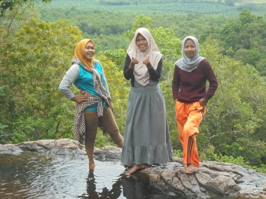 Three women stand on a rocky surface near water, overlooking a lush green landscape. They are dressed in casual attire with headscarves, and they appear happy and relaxed in their natural surroundings.