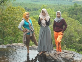 Three women stand on a rocky surface near water, overlooking a lush green landscape. They are dressed in casual attire with headscarves, and they appear happy and relaxed in their natural surroundings.