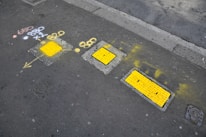 Several rectangular yellow utility covers are embedded in a sidewalk with surrounding marks made in yellow spray paint, including arrows and symbols. Additional figures and symbols are painted in white and orange. The sidewalk is gray, and there is a road surface adjacent to it.