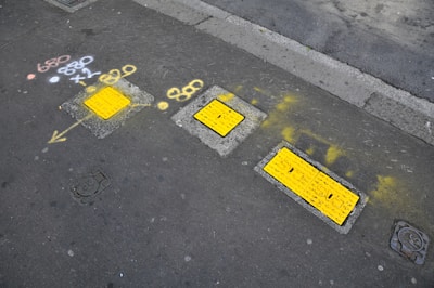 Several rectangular yellow utility covers are embedded in a sidewalk with surrounding marks made in yellow spray paint, including arrows and symbols. Additional figures and symbols are painted in white and orange. The sidewalk is gray, and there is a road surface adjacent to it.