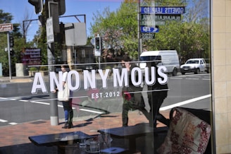 A glass window reflects a street view, with the word 'ANONYMOUS' and 'EST. 2012' prominently displayed. People are seen walking past, and cars are parked or driving by. The reflection shows trees and street signs.
