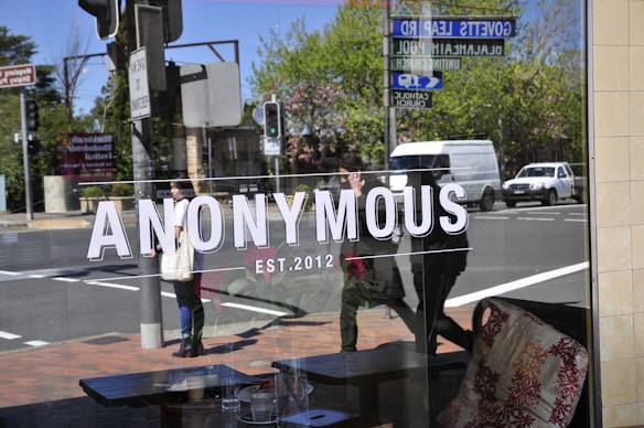 A glass window reflects a street view, with the word 'ANONYMOUS' and 'EST. 2012' prominently displayed. People are seen walking past, and cars are parked or driving by. The reflection shows trees and street signs.