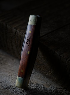 A wooden-handled tool or object with metallic ends stands upright against a textured, wooden surface. The handle has brass-colored rivets and an engraved design resembling an animal's head.