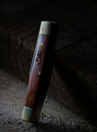 A wooden-handled tool or object with metallic ends stands upright against a textured, wooden surface. The handle has brass-colored rivets and an engraved design resembling an animal's head.