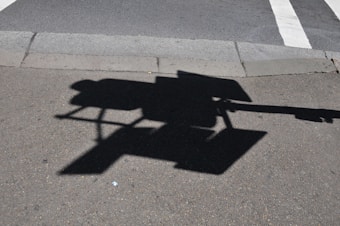A shadow of a traffic camera or surveillance device is cast on an asphalt road near a sidewalk and a crosswalk with white stripes.