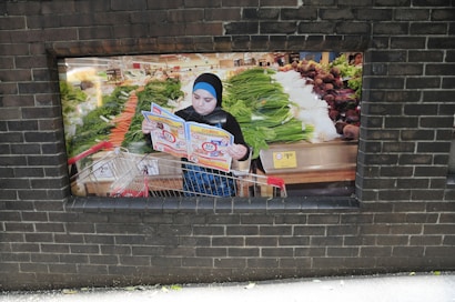 A person stands with a shopping cart in a grocery store, reading a colorful brochure. The store features an array of fresh produce, including leafy greens and root vegetables displayed on shelves. The atmosphere is busy and colorful, indicating a bustling market environment.