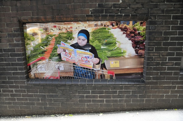 A person stands with a shopping cart in a grocery store, reading a colorful brochure. The store features an array of fresh produce, including leafy greens and root vegetables displayed on shelves. The atmosphere is busy and colorful, indicating a bustling market environment.