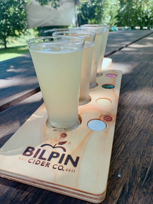 A wooden flight board holding several glasses filled with a light-colored beverage is set on a rustic wooden table. Sunlight filters through surrounding greenery, creating a relaxed outdoor setting. The board prominently features the logo 'BILPIN CIDER CO.'