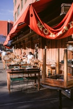 An outdoor restaurant seating area with multiple wooden tables and chairs arranged along the sidewalk. Red awnings with gold lettering provide shade. A few people are seated at some of the tables, suggesting a relaxed dining atmosphere. The building's exterior features a rustic wooden facade.
