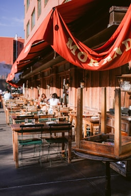 An outdoor restaurant seating area with multiple wooden tables and chairs arranged along the sidewalk. Red awnings with gold lettering provide shade. A few people are seated at some of the tables, suggesting a relaxed dining atmosphere. The building's exterior features a rustic wooden facade.