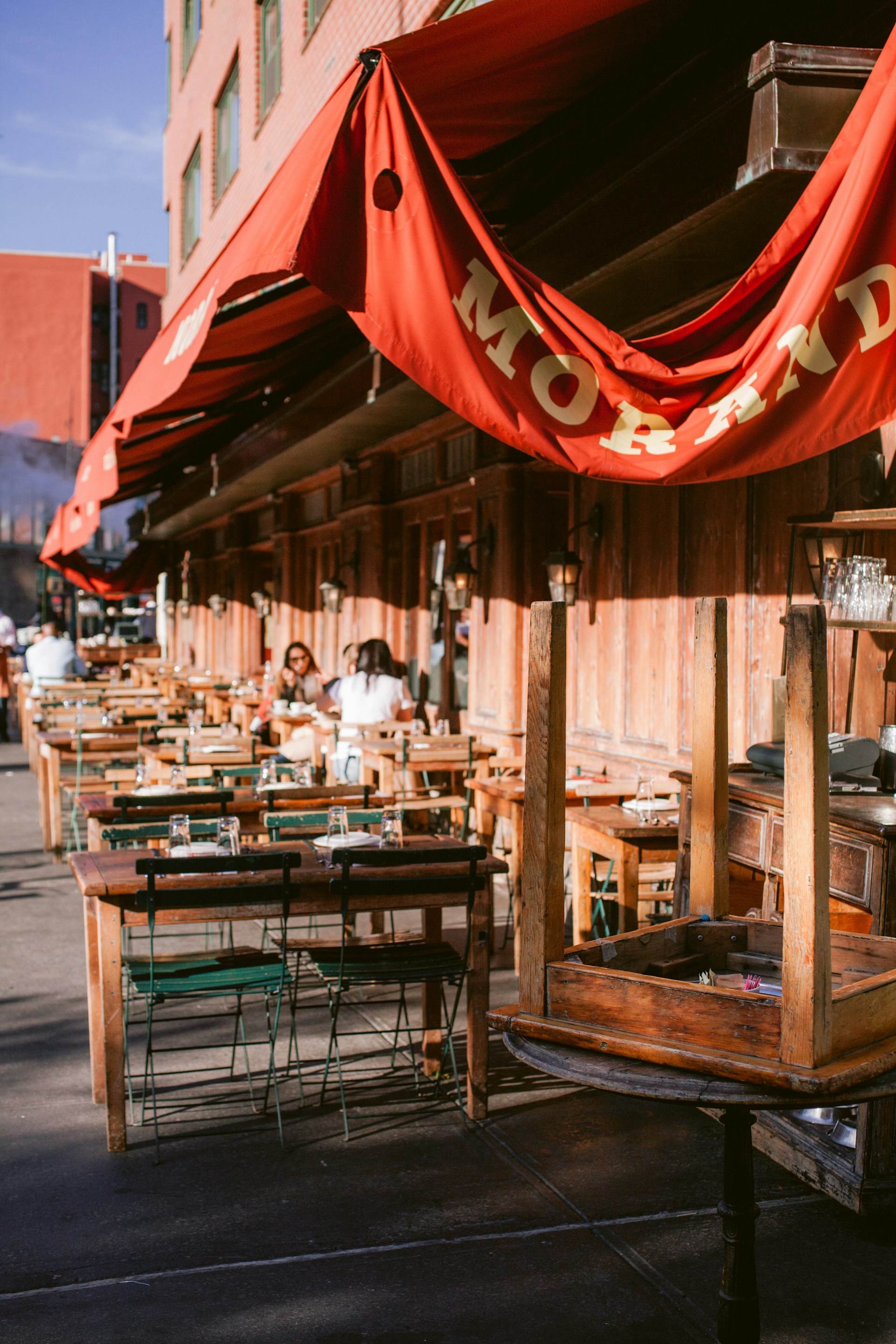 An outdoor restaurant seating area with multiple wooden tables and chairs arranged along the sidewalk. Red awnings with gold lettering provide shade. A few people are seated at some of the tables, suggesting a relaxed dining atmosphere. The building's exterior features a rustic wooden facade.