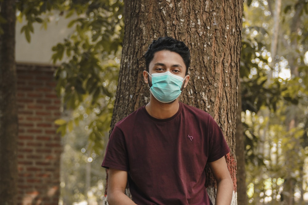 man in maroon crew neck t-shirt with face mask standing beside brown tree during daytime