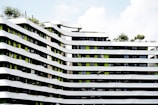 A modern architectural building featuring undulating white and green facades with multiple floors. The building's design includes a pattern of curved lines with greenery present on the rooftop areas. Several windows align in between the white strips, adding a contemporary urban feel to the structure.