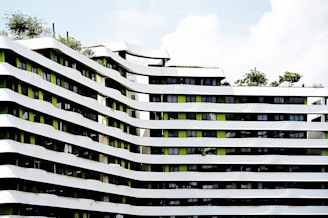 A modern architectural building featuring undulating white and green facades with multiple floors. The building's design includes a pattern of curved lines with greenery present on the rooftop areas. Several windows align in between the white strips, adding a contemporary urban feel to the structure.