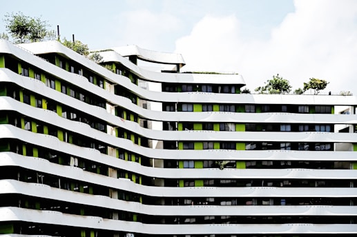 A modern architectural building featuring undulating white and green facades with multiple floors. The building's design includes a pattern of curved lines with greenery present on the rooftop areas. Several windows align in between the white strips, adding a contemporary urban feel to the structure.