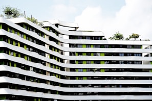 A modern architectural building featuring undulating white and green facades with multiple floors. The building's design includes a pattern of curved lines with greenery present on the rooftop areas. Several windows align in between the white strips, adding a contemporary urban feel to the structure.