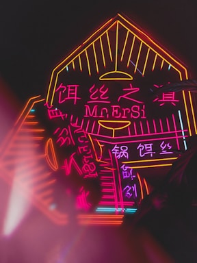 Brightly lit neon sign featuring vibrant pink and orange colors with both English and Chinese characters. The design includes angular and geometric elements framing the text.