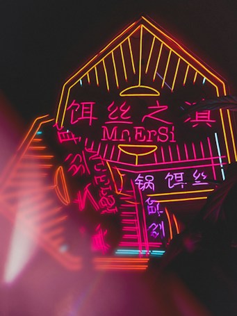 Brightly lit neon sign featuring vibrant pink and orange colors with both English and Chinese characters. The design includes angular and geometric elements framing the text.