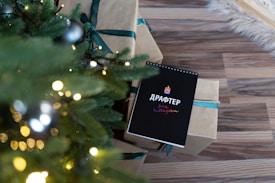 A festive setting with a decorated Christmas tree featuring lights and ornaments. Next to the tree, there are wrapped gifts with green ribbon and a black notebook on top of the presents displaying some colorful text.