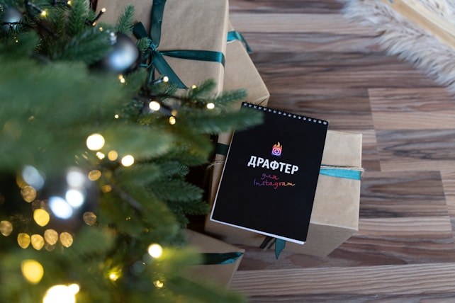 A festive setting with a decorated Christmas tree featuring lights and ornaments. Next to the tree, there are wrapped gifts with green ribbon and a black notebook on top of the presents displaying some colorful text.