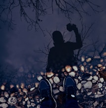 A reflection in water shows a person holding a camera, surrounded by bare tree branches. Pebbles line the edge of the water where two shoes are visible, giving the appearance of someone standing at the water's edge.