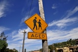 A yellow traffic sign mounted on a pole against a clear blue sky. The sign features an image of two figures walking and the word 'AGED' beneath it. In the background, there are trees, utility poles, and rooftops.