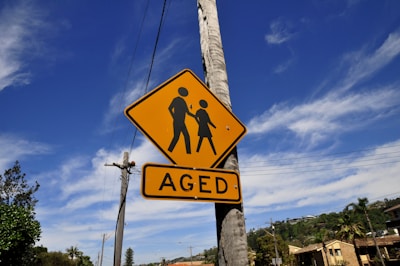 A yellow traffic sign mounted on a pole against a clear blue sky. The sign features an image of two figures walking and the word 'AGED' beneath it. In the background, there are trees, utility poles, and rooftops.