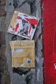A textured surface featuring two layered square artworks. The upper piece contains a collage with abstract red and white paint and a black and white photograph of a person. The lower piece consists of a grid made from strips of yellow tape or paper, revealing a small colored object beneath. The background is composed of a rough grey wall with different textures, crackling paint, and a broad red stripe on the right.