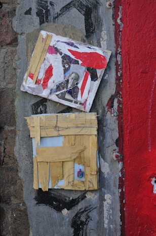 A textured surface featuring two layered square artworks. The upper piece contains a collage with abstract red and white paint and a black and white photograph of a person. The lower piece consists of a grid made from strips of yellow tape or paper, revealing a small colored object beneath. The background is composed of a rough grey wall with different textures, crackling paint, and a broad red stripe on the right.