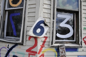 A weathered white wooden house exterior features large, colorful numbers painted and stuck on it. The numbers include a blue 7 on a window, a white 3 on another window, and a slightly crumpled metal sign with the number 6 affixed to the wall. Additional numbers 4, 2, and some others are painted in various colors like red, blue, and purple on the siding.