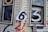 A weathered white wooden house exterior features large, colorful numbers painted and stuck on it. The numbers include a blue 7 on a window, a white 3 on another window, and a slightly crumpled metal sign with the number 6 affixed to the wall. Additional numbers 4, 2, and some others are painted in various colors like red, blue, and purple on the siding.