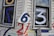 A weathered white wooden house exterior features large, colorful numbers painted and stuck on it. The numbers include a blue 7 on a window, a white 3 on another window, and a slightly crumpled metal sign with the number 6 affixed to the wall. Additional numbers 4, 2, and some others are painted in various colors like red, blue, and purple on the siding.
