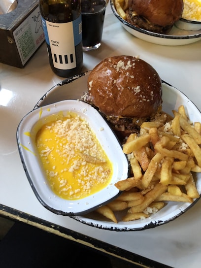 A plate of food featuring a burger in a bun topped with shredded cheese, a serving of golden French fries, and a container of yellow cheese sauce sprinkled with more shredded cheese. There is a bottle of 'Coco Milk' stout beer and a glass of dark drink in the background.