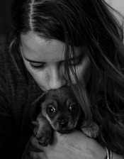 A person with long hair gently holds a small, wide-eyed puppy close to their face in an affectionate manner. The image is in black and white, emphasizing the emotional connection.