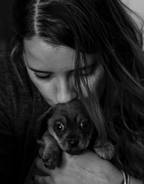 A person with long hair gently holds a small, wide-eyed puppy close to their face in an affectionate manner. The image is in black and white, emphasizing the emotional connection.