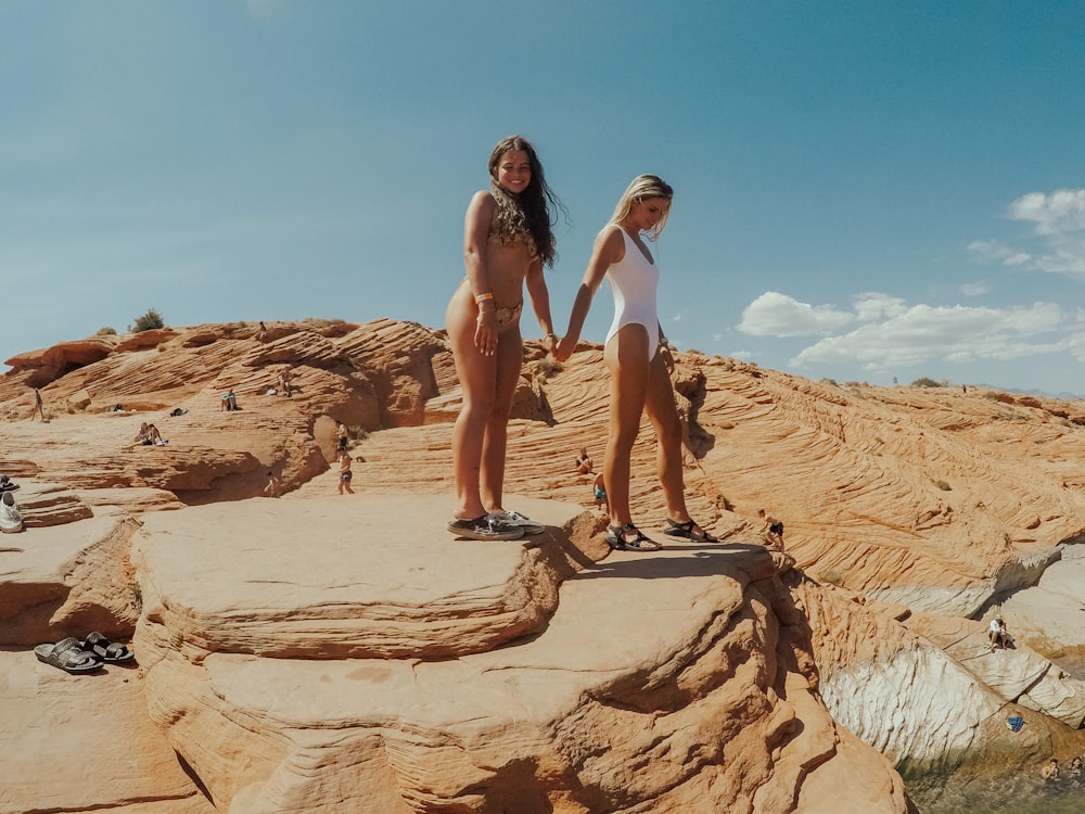 2 women in white tank top and white panty standing on brown rock formation during daytime